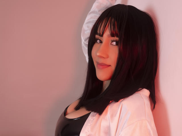 CelesteRusso with black hair, bangs, and a white shirt, leaning against a wall, wearing a black leather top, showing her tits, and smiling slightly.