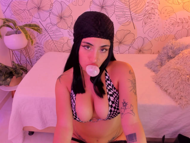DiynaStar with black beanie, bubble gum in mouth, wearing checkered bikini top, showing big tits, tattoos on arm, sitting on bed, purple and pink lighting, looking at camera.