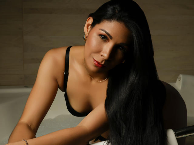 EliseBell has long black hair, tan skin, and small tits, leaning in a bathtub wearing a black bra, with a subtle smile and red lipstick, looking at the camera.