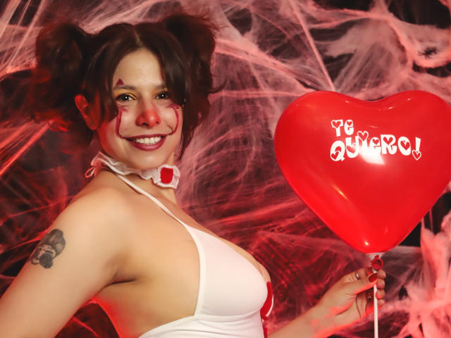 SarahWelinton with puffy brown hair in double buns, white halter top, red heart balloon, facial paint, smiling, tattoo on right arm, holding balloon, pink spider web background.