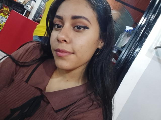 MarilynHills with medium brown skin, long black hair, and winged eyeliner, wearing a brown ribbed shirt, smiling softly, showing small black earrings and a slight pimple on her cheek.