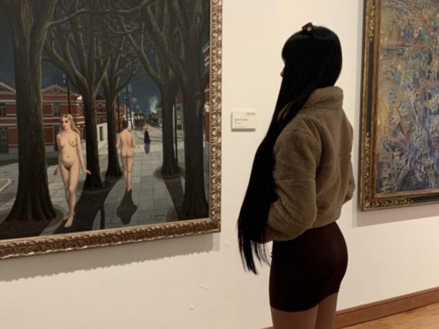 ThaliaCollins with long black hair, brown sweater, and tight brown shorts, stands before a painting of two naked women with small tits, standing in a dark, tree-lined street.
