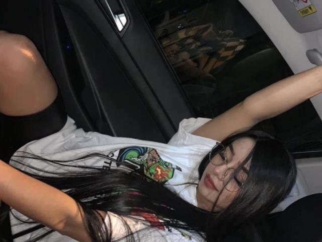 Thalia Collins, mit langen schwarzen Haaren, trägt ein weißes T-Shirt und schwarze Shorts und liegt in einem Autositz, die Arme erhoben, und schaut mit einem leichten Lächeln in die Kamera..