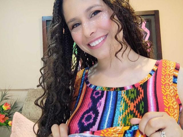 SarahWelinton with curly brown hair, sparkling glitter on her cheeks, smiling, wearing a colorful, patterned dress, holding her dress up, showing her tits, in a well-lit room.