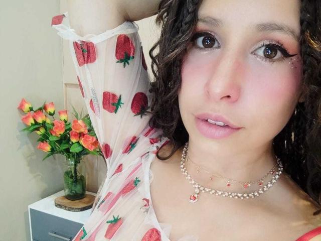 SarahWelinton with curly brown hair, pink lipstick, and a strawberry-patterned sheer top, wearing a pearl necklace, arm raised, pink blush, and orange roses in a vase beside her.