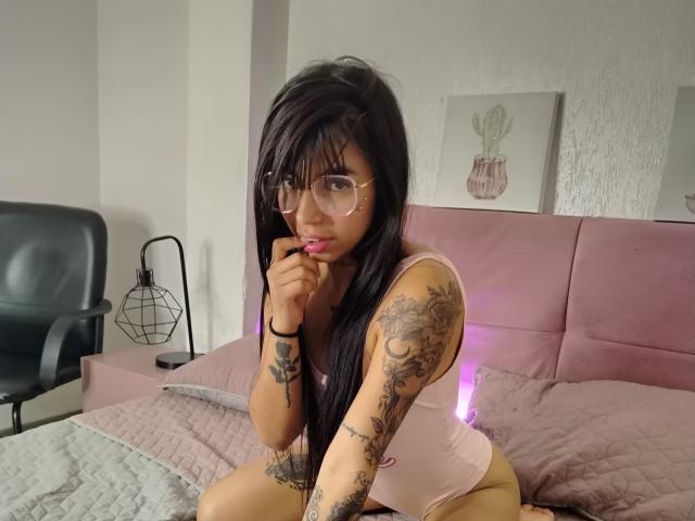 KendallPrakth with long black hair, glasses, pink tank top, tattoos, and pink lips, biting finger, kneeling on bed with pink bedspread, purple light, and black chair in background.