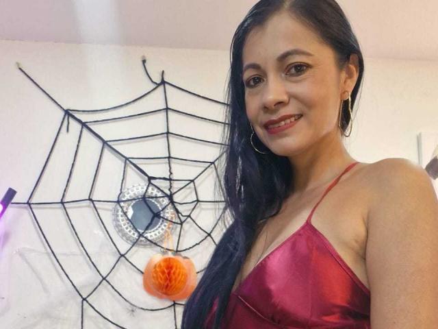 Melissa Baud mit langem, schwarzem Haar, rotem Seidenkleid, Reifenohrringen, lächelnd, wobei ihre Brüste sichtbar sind, ein kleiner, orangefarbener Pompon hängt hinter ihr, schwarze Spinnennetz-Wanddekoration..