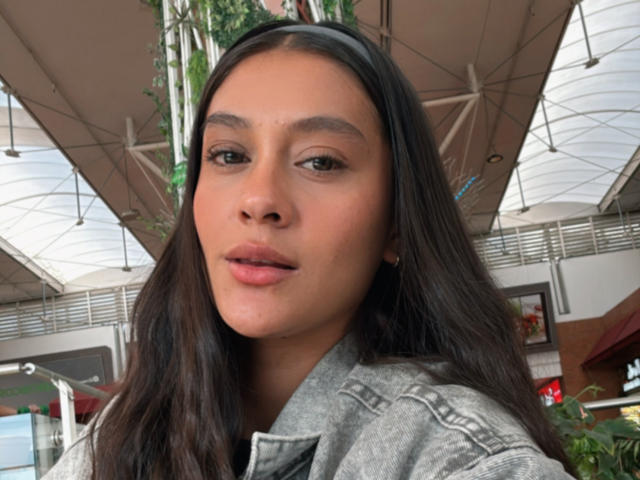 ThaliaAres has brown skin, long dark hair, and a black headband. She's wearing a gray denim jacket, looking at the camera with slightly parted pink lips.