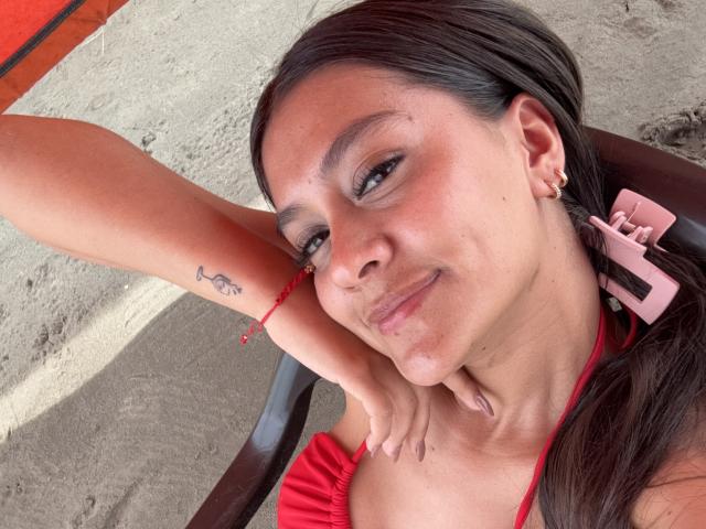 ThaliaAres with dark hair, red halter top, and pink hairclip, leaning on chair, smiling, showing small tattoo on upper arm, red bracelet, and gold earrings.