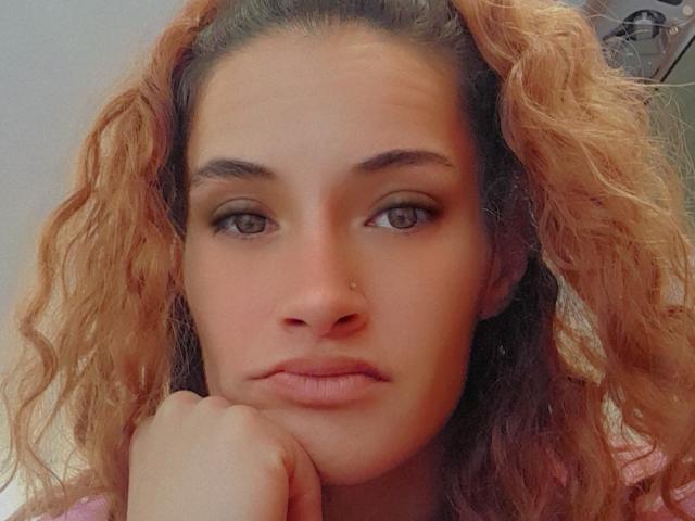 TheQueEnoFDabz has curly, orange-brown hair, a nose ring, and brown eyes with subtle makeup. She's resting her chin on her hand, looking serious.