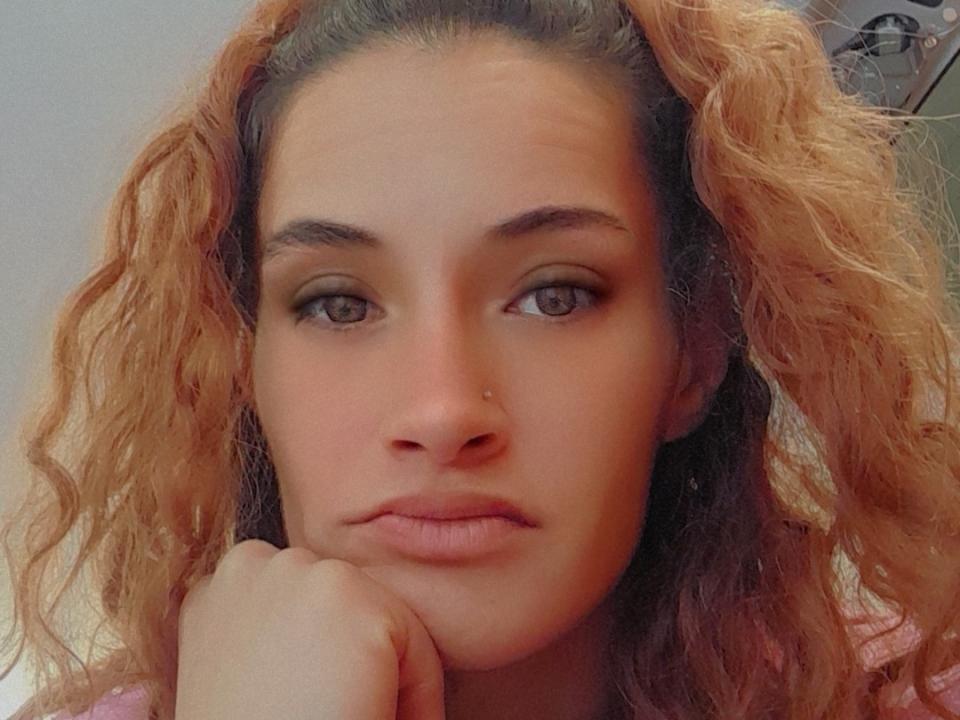 TheQueEnoFDabz has curly, orange-brown hair, a nose ring, and brown eyes with subtle makeup. She's resting her chin on her hand, looking serious.