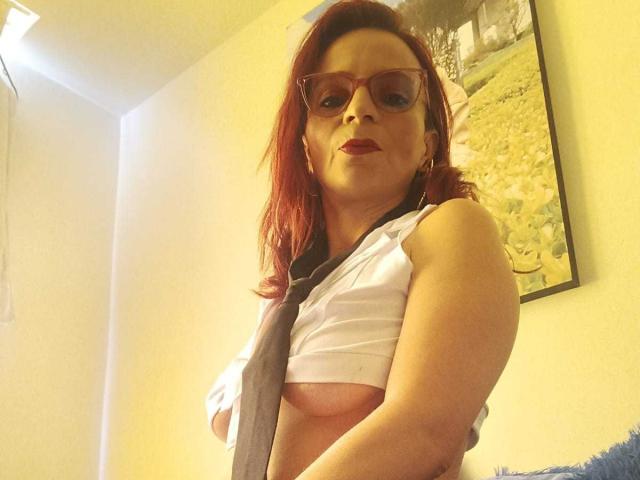 MissEllien with red hair, glasses, and red lipstick, wearing a white shirt, grey tie, and no bra, exposing her tits. She's looking down, slightly smiling.