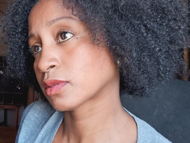 Close-up of Lizane6. She has medium brown skin, brown eyes, and curly black hair. Her face is slightly turned to the right, showing a serious expression with pursed lips. She wears a light blue, V-neck shirt. Her nose is small and she has a small black mole on her right cheek. The texture of her curly hair is detailed and her eyebrows are thin and slightly raised. The image focuses on her upper face and shoulders.