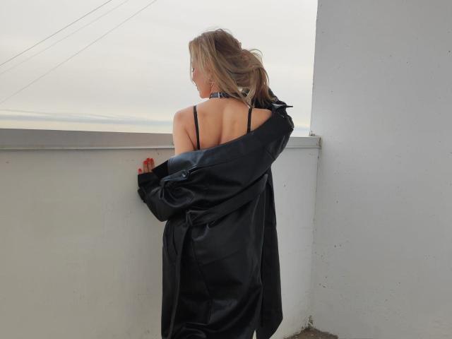SweetLadyXX with her back to the camera, wearing a black satin dress and slipping off a black jacket, showing bare shoulders and a black strap.