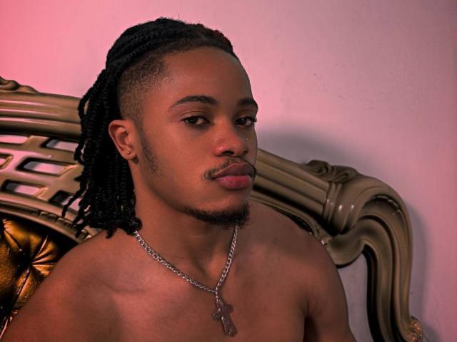Kobalad, com pele escura, dreadlocks pretos e uma pequena barba, sem camisa, usando uma corrente de prata com uma placa de identificação, sentado em uma cadeira dourada e ornamentada..