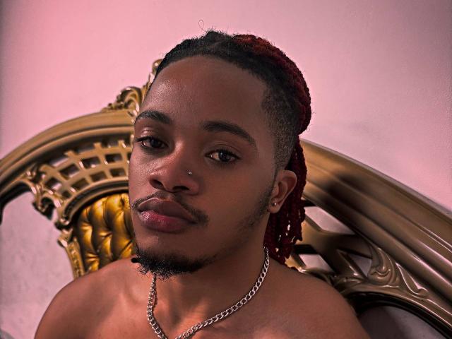 Kobalad sem camisa, com pele escura, cabelo trançado vermelho, piercing pequeno no nariz, colar de corrente e barba, olhando ligeiramente para o lado, sentado em uma elaborada cabeceira dourada..