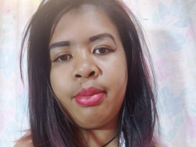 MariahSquirting69 has dark, straight hair, brown eyes, and pink lipstick. Her eyebrows are thick, and she has a slight, closed-lip smile. Her skin is medium brown.
