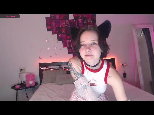  OliviaFlex has short, dark brown hair and wears black furry cat ears. She's smiling at the camera, leaning slightly forward. She's dressed in a white tank top with red trim and a cherry design, paired with pink and white patterned pajamas. She has tattoos on her right arm. The room has a bed with beige pillows and a pink heart-shaped pillow on a small black table. A wall is decorated with a grid of pink photos. The lighting is soft with pink and white tones. The image has a watermark in the top left corner.