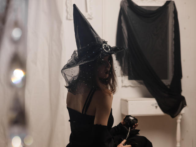 MarieLoving wears a black witch hat with a sheer, polka-dotted veil. She has dark, wavy hair and a black off-shoulder dress. She's seated, looking back over her shoulder with a subtle, playful expression. In the background, a white wall with a draped black cloth is visible. The image uses soft, diffused lighting, emphasizing the dark, mystical theme. The composition centers MarieLoving, creating a focused, slightly mysterious atmosphere.