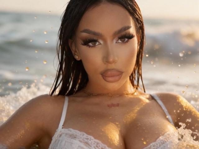 SierraDeNoir with wet brown hair, full eyelashes, and tan skin, wearing a white lace top, her big tits glistening with water, a small tattoo above her cleavage.