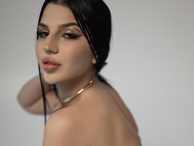 KamilaVibes with wet black hair, glowing skin, and subtle makeup, showing her bare shoulders and a gold necklace, looking back with a slight smile, her pink lips prominent.