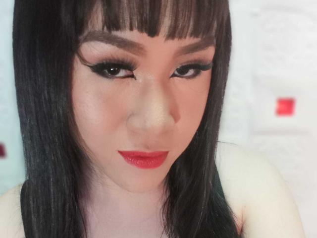 CockZilLaHotTs, con capelli neri lisci, frangia e rossetto rosso, ha un eyeliner scuro e rossetto rosso, pelle chiara, guarda in basso con un leggero sorriso, indossando una maglietta nera..