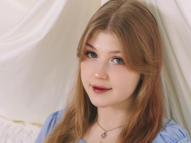 StellaLane has light brown, wavy hair, green eyes, and a cute smile. She's wearing a blue top with a silver necklace and has shiny pink lips.