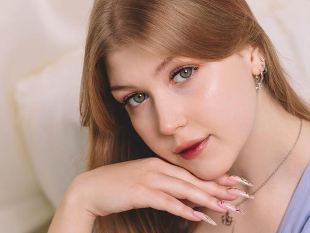 StellaLane mit hellbraunen Haaren, grünen Augen und rosafarbenen Lippen, die ihr Kinn in die Hand legt, wobei ihre langen, gepflegten Nägel zu sehen sind. Sie trägt eine Halskette und hat ein leichtes Lächeln..