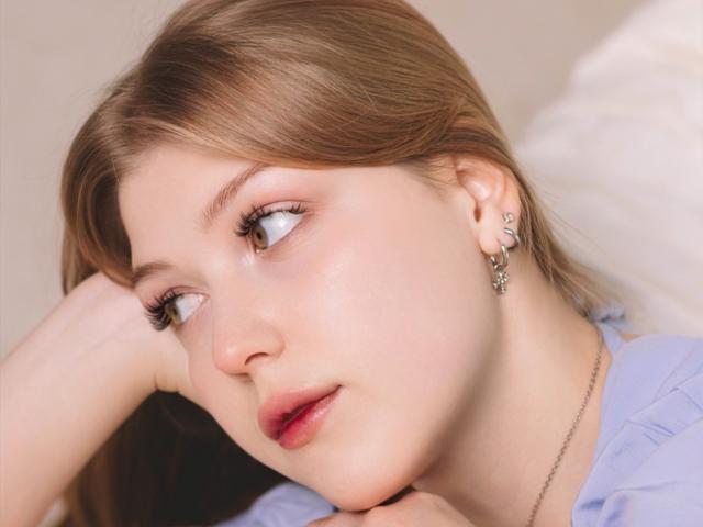 StellaLane's face, showing pale skin, red lips, and brown hair, with a side profile, hand on cheek, looking up, wearing a blue shirt, and silver earrings.