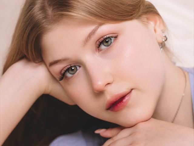 StellaLane has light green eyes, fair skin, and pink lips, with her brown hair down, resting her head on her arm, wearing a silver earring, looking slightly to the side.