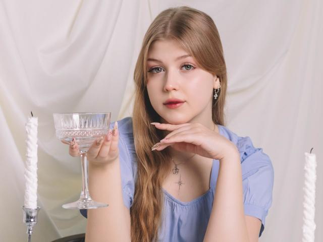 StellaLane with long blonde hair, blue eyes, and light makeup, holding a glass goblet, wearing a blue dress, and resting her chin on her hand, with white candles.