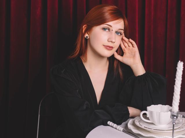 PollyFoxy with red hair, black dress, ear gauges, hand on cheek, looking at camera, white cup and saucer on table, red velvet curtain in background.