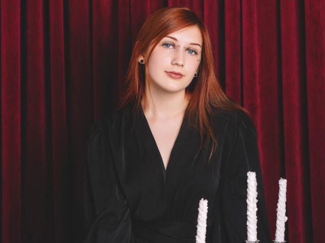 PollyFoxy has red hair, pale skin, and small earrings, wearing a black velvet robe with white trim, looking slightly to the left with a calm expression.