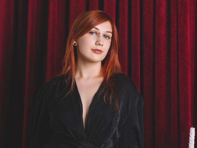 PollyFoxy with red hair, blue eyes, and black robe, showing a hint of her tits, standing in front of red velvet curtains, looking up with a slight smile.