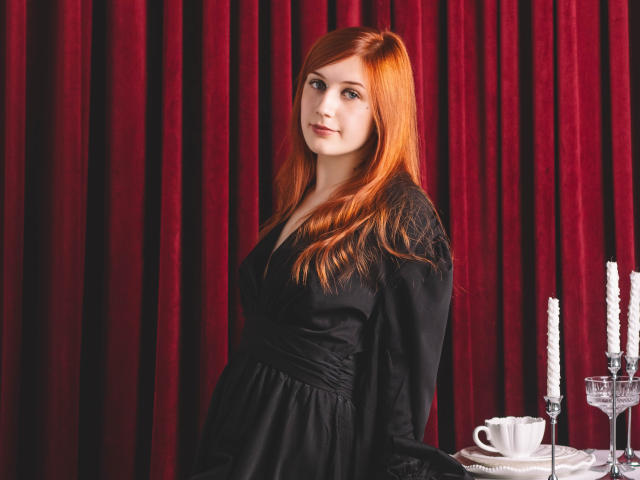 PollyFoxy with red hair, wearing a black dress, standing by a table with white candles and a cup. She's got a cute face and a sexy, attractive look.
