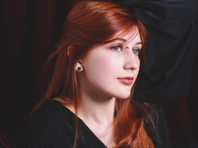 PollyFoxy has red hair, blue eyes, black ear studs, and a small nose ring. She's wearing a black shirt, looking to the side with a slight smile.