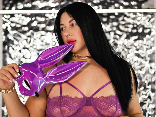 ArianaDarko with black hair, purple lace bra, and red lipstick, holding a purple bunny mask, her red lips near the mask's ear, wearing a gold bracelet.
