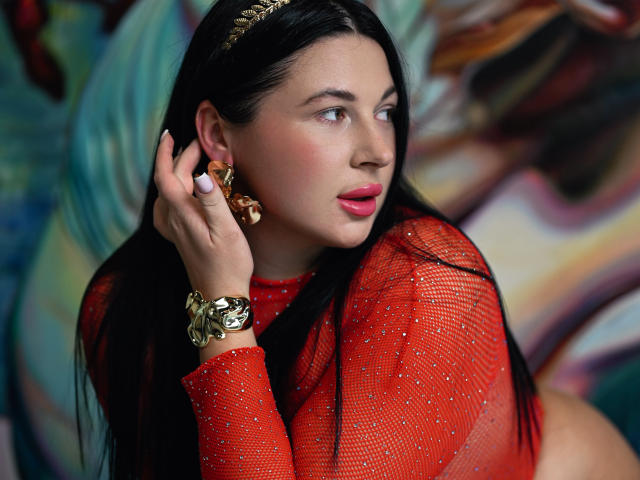 ArianaDarko with long black hair, red mesh top, gold earrings, and gold bracelet, holding ear, looking sideways, pink lipstick, serious expression, soft light. Boobs visible under top.