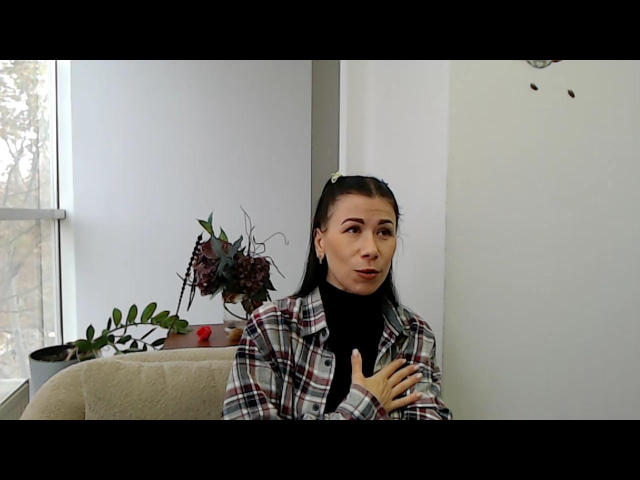 Glidia with dark hair in a ponytail, wearing a plaid shirt over a black turtleneck, has her hand near her mouth, looking slightly to the right. Potted plants in background.