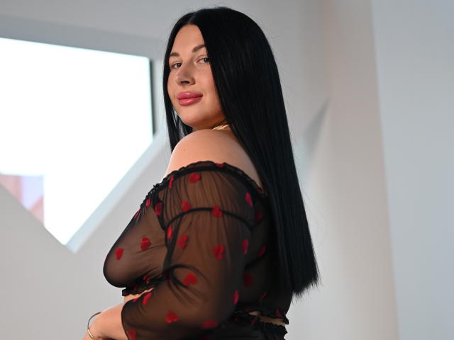 ArianaDarko with long black hair, wearing a sheer black top with red rose patterns, showing her bare shoulders and smiling at the camera, standing in a bright room.