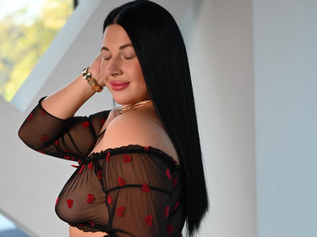ArianaDarko with long black hair, wearing a sheer black top with red hearts, smiling, eyes closed, hand on head, gold bracelet, and gold necklace, showing her tits.
