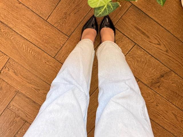 KaiaLein's legs in light blue pants with black pointed shoes on a wooden floor, a green leaf partially in the top right corner.