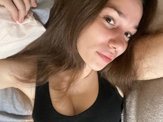 KaiaLein allongée sur un lit, cheveux bruns, peau claire, débardeur noir, petites seins, léger sourire, yeux marrons, bras gauche levé, bras droit reposant sur l'oreiller, détendue..