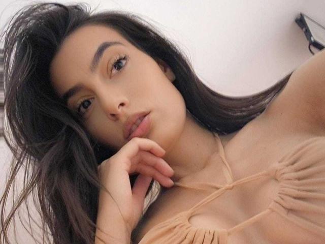 NinaGomez with long dark hair, light brown skin, and small tits, wearing a beige strappy top, hand near mouth, looking at camera, slightly parted lips, subtle makeup.