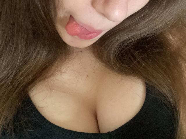 KaiaLein's face and upper chest with her tongue slightly out, pink lips, brown wavy hair, black top, showing her cleavage and small nipples.