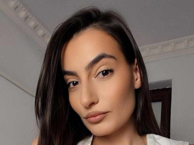 Close-up of NinaGomez with a neutral expression. She has medium brown skin, straight dark brown hair parted to the side, and thick, well-groomed eyebrows. Her brown eyes are slightly downcast, and she has subtle, natural-looking makeup with a light pinkish-nude lip. She wears a white top, visible at the bottom of the frame. The background is a blurred, simple interior with a dark wooden door and a white ceiling with a decorative border. The image has a soft, realistic texture.