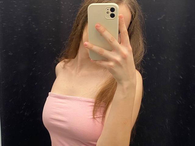 KaiaLein with long brown hair, holding a white iPhone, covering her face, wearing a pink strapless top, showing her tits, against a dark background.