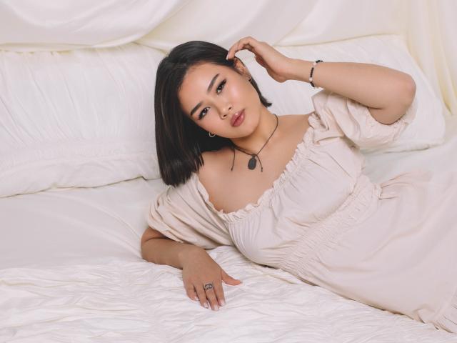 ArianaFlower, a young Asian woman with straight black hair, lies on a white bed. She wears a beige off-shoulder dress with puffed sleeves and a black necklace with a small pendant. Her right hand touches her hair, and her left hand rests on the bed. She has a ring on her left middle finger. The composition is soft and relaxed, with a focus on her calm expression and delicate features. The texture of the bed and dress is slightly crumpled, adding depth to the image.