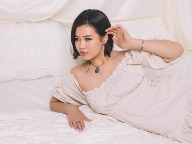 ArianaFlower has short black hair, wearing an off-shoulder beige dress, lying on white pillows and bedding. She's touching her ear with a bracelet on her wrist.