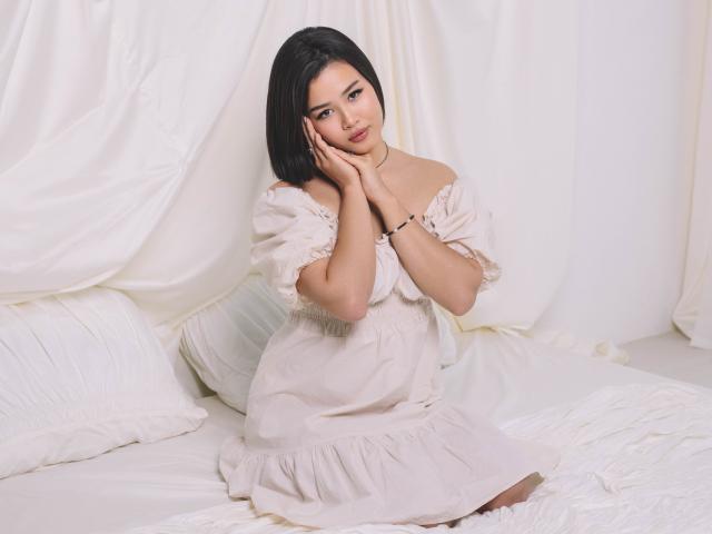 ArianaFlower with black hair, wearing an off-shoulder white dress, sitting on a bed, touching her face, looking at the camera, white pillows and sheets in the background.